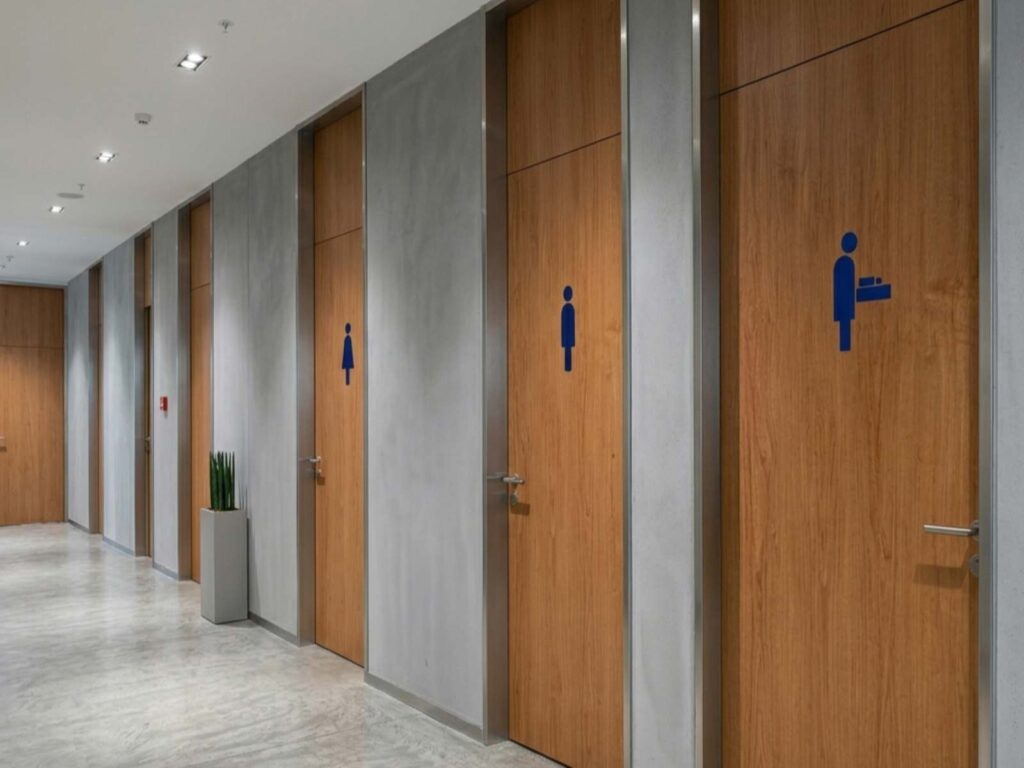 Dunkelblaue Symbole für die Herren- und Damen-Toiletten sowie für einen Wickelraum auf Holztüren in einem Sanitärraum.
