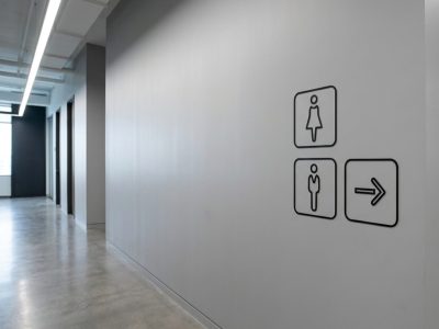 Schwarz eingerahmte Symbole für die Herren- und Damen-Toiletten mit einem nach rechts weisenden Pfeil in einem Flur.